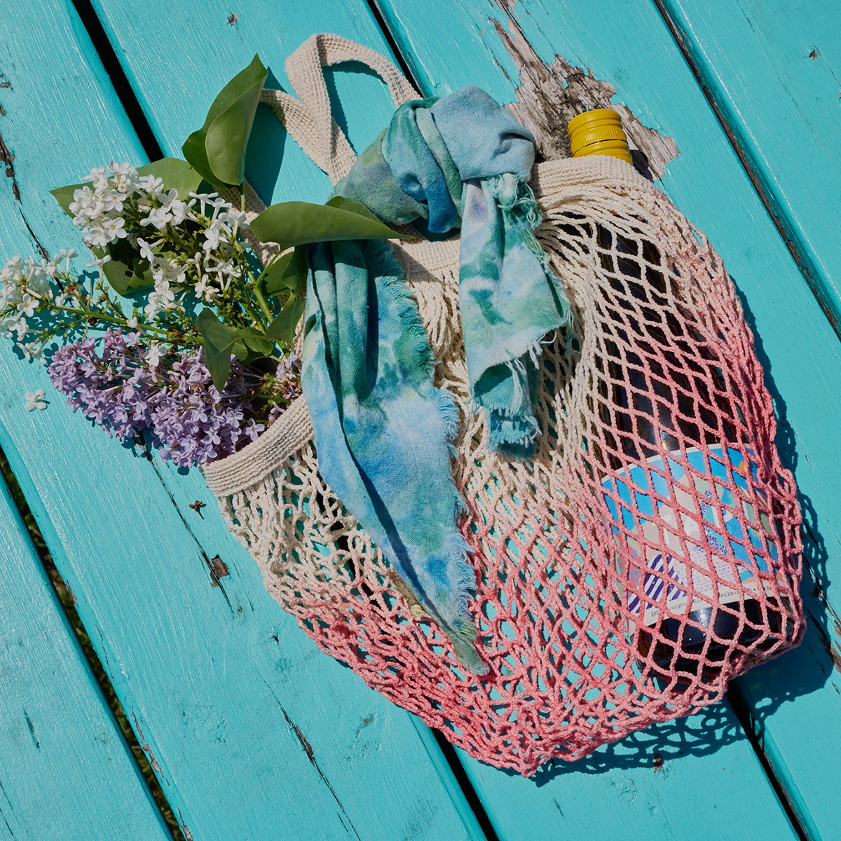 Colorful mesh bag with a bottle, bandana, and flowers on a turquoise wooden surface