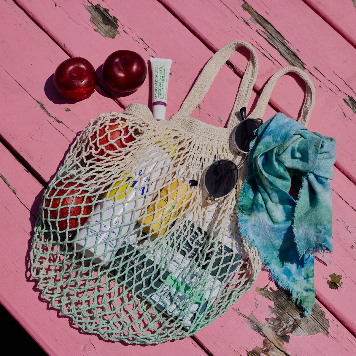 Mesh bag with fruits, tube of cream, and sunglasses on a pink wooden surface