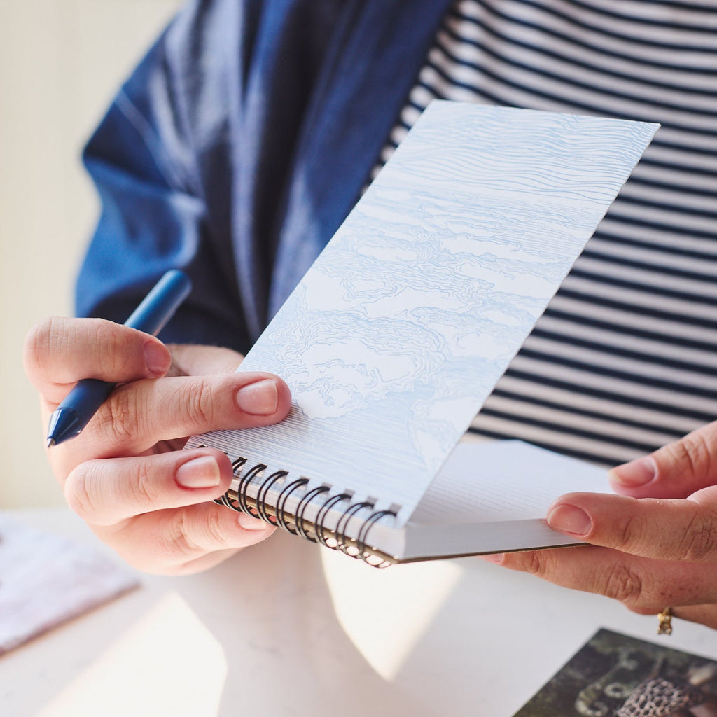 A close-up of a person holding an open spiral-bound notebook with a blue pen, wearing a navy and white striped shirt and a blue cardigan.