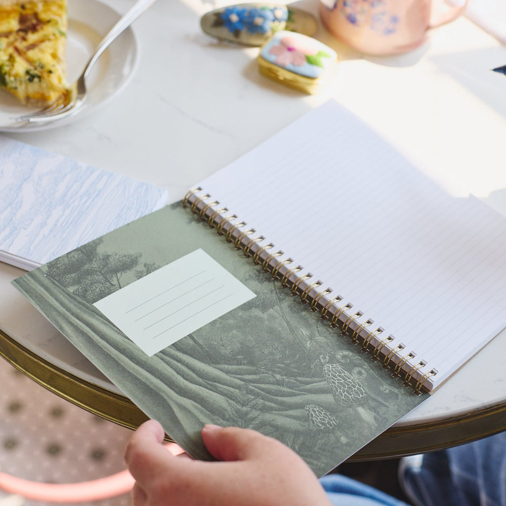 A person's hands holding an open lined spiral notebook with a green patterned cover, sitting at a round white table with a blue pen in the other hand. A plate of food and a mug are visible in the background.