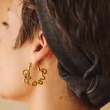 A close-up side profile of a person wearing a gold-toned, abstract design hoop earring with small, multicolored gemstone accents, paired with a vintage black denim fabric headband.