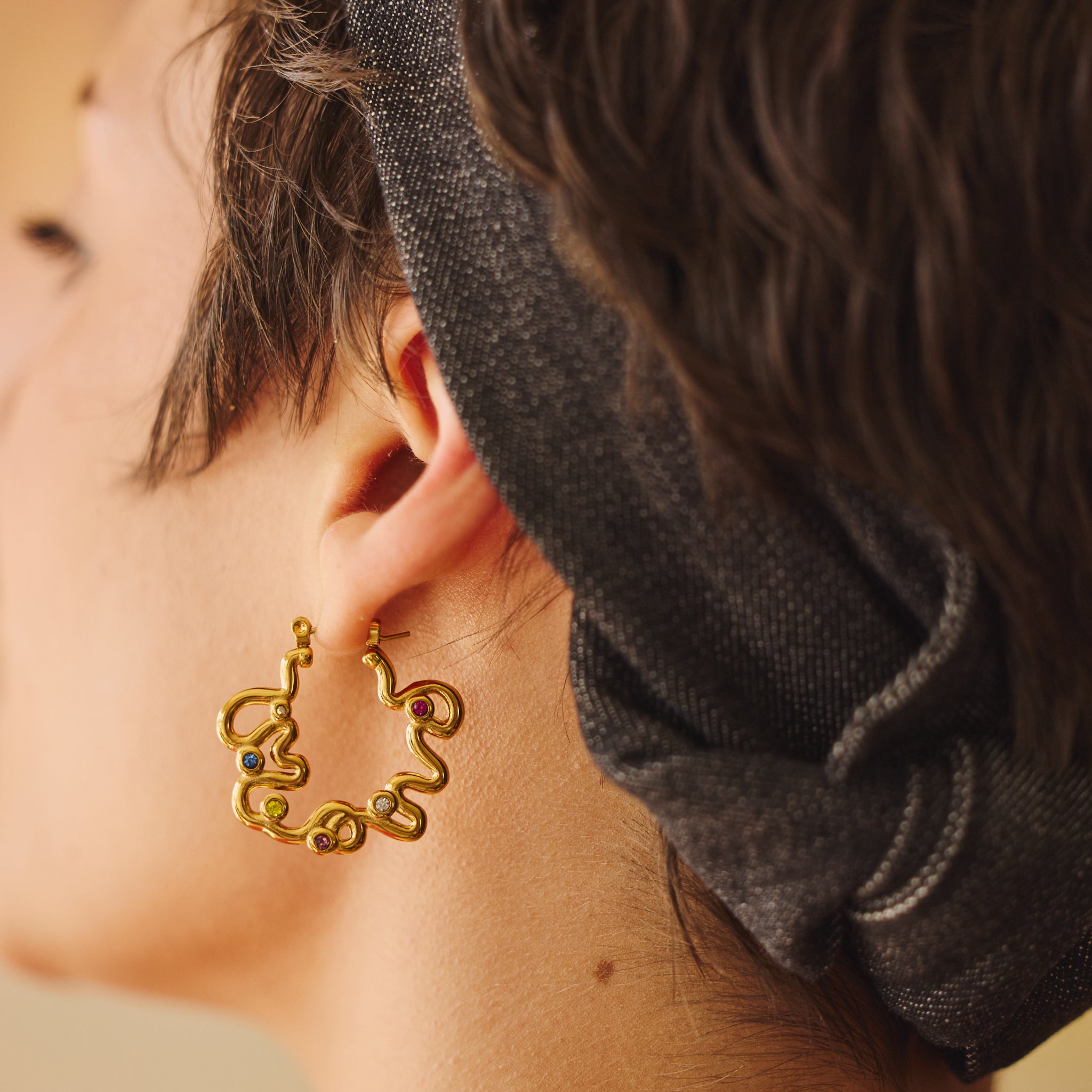 A close-up side profile of a person wearing a gold-toned, abstract design hoop earring with small, multicolored gemstone accents, paired with a vintage black denim fabric headband.