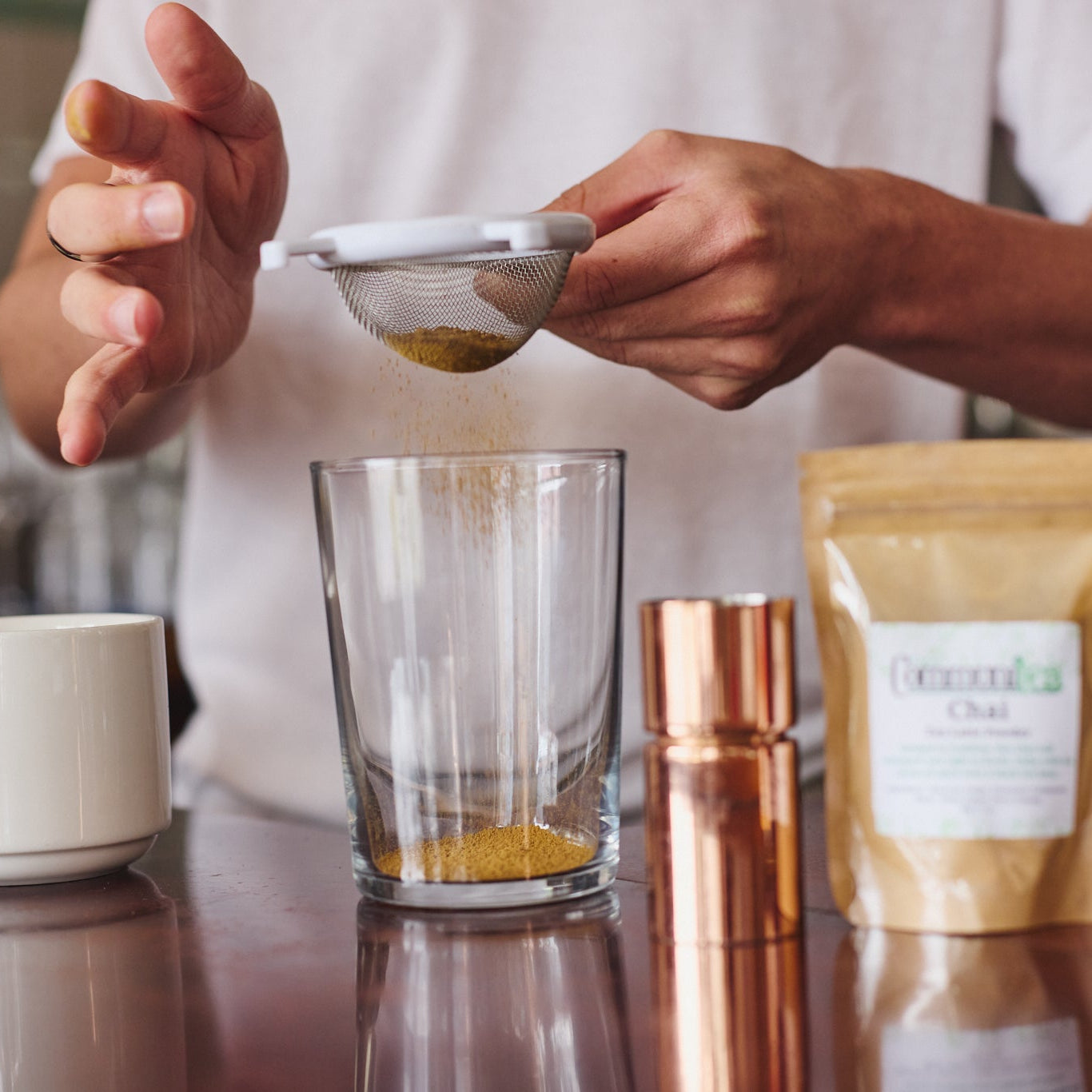 A person in a white shirt holds a small metal sieve above a clear glass container, pouring yellow powder into it. On the counter are a white mug, a copper shaker, a pouch labeled 'Herbal Chai', and a bottle of syrup in a warm indoor setting.