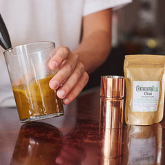 A person using an electric milk frother to create foam in a glass of tea on a dark wooden table, with a white mug, a copper shaker, and a bag of Chai tea nearby.