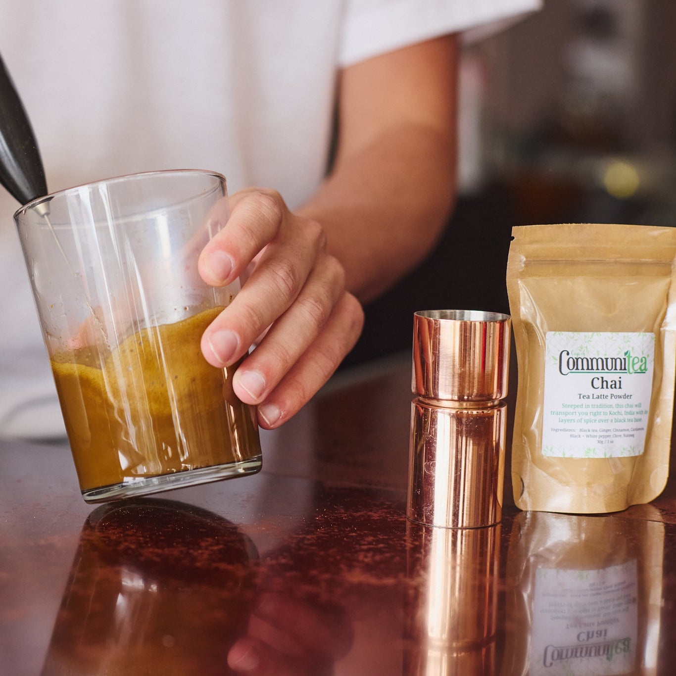 A person using an electric milk frother to create foam in a glass of tea on a dark wooden table, with a white mug, a copper shaker, and a bag of Chai tea nearby.