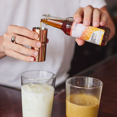 A person pours a dark amber liquid from a bottle labeled 'Chai' into a copper jigger, with two glasses below—one filled with white liquid and one with an amber drink on a dark wooden table.