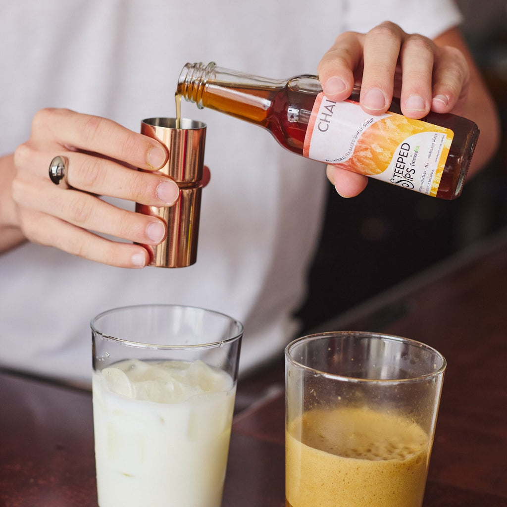A person pours a dark amber liquid from a bottle labeled 'Chai' into a copper jigger, with two glasses below—one filled with white liquid and one with an amber drink on a dark wooden table.