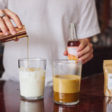 A person in a white shirt pours a golden liquid from a copper jigger into a glass of milk, while holding a small bottle in a bar setting. Another glass with an amber drink sits beside it on a dark wooden counter.
