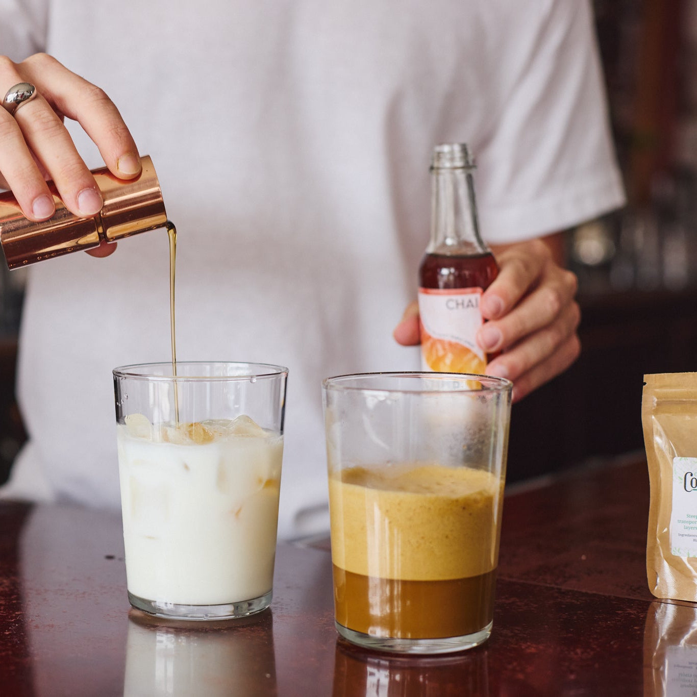 A person in a white shirt pours a golden liquid from a copper jigger into a glass of milk, while holding a small bottle in a bar setting. Another glass with an amber drink sits beside it on a dark wooden counter.
