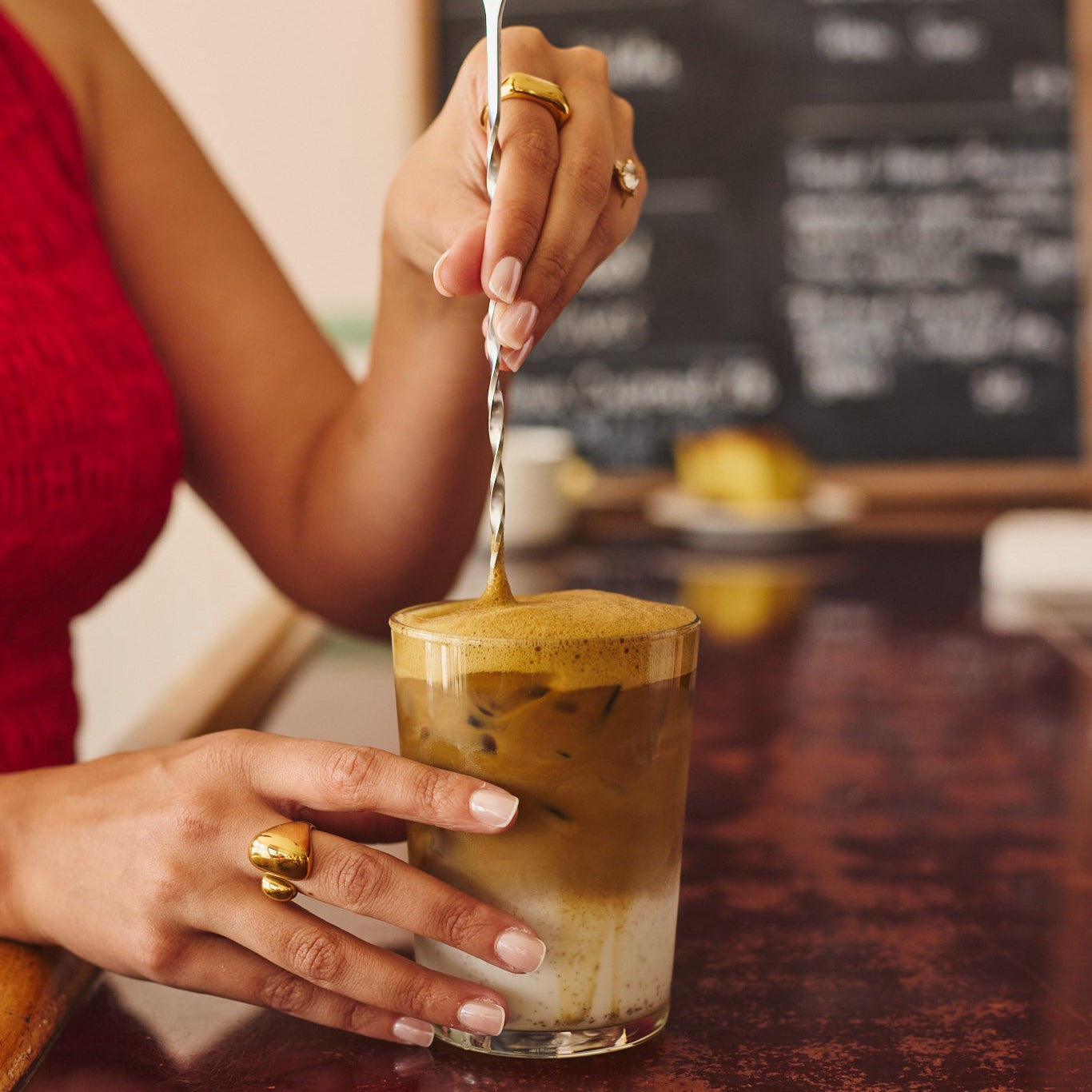 A person in a red sleeveless top stirs a layered iced tea latte in a clear glass with a twisted spoon on a dark reddish-brown counter, with a blurred cafe chalkboard in the background.
