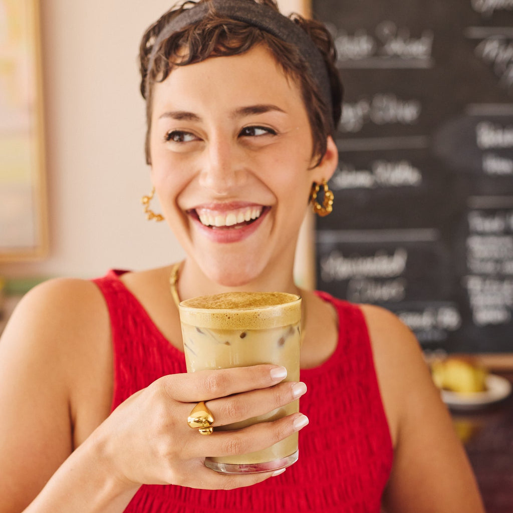 A woman in a red sleeveless dress holding an iced tea latte in a cafe setting, wearing gold jewelry.