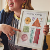 A woman in a navy cardigan and striped shirt holds up a white-framed art print titled 'Shapes of Buffalo' while pointing at it. The print features colorful hand-drawn illustrations of shapes and patterns using Buffalo landmarks.
