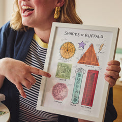 A woman in a navy cardigan and striped shirt holds up a white-framed art print titled 'Shapes of Buffalo' while pointing at it. The print features colorful hand-drawn illustrations of shapes and patterns using Buffalo landmarks.