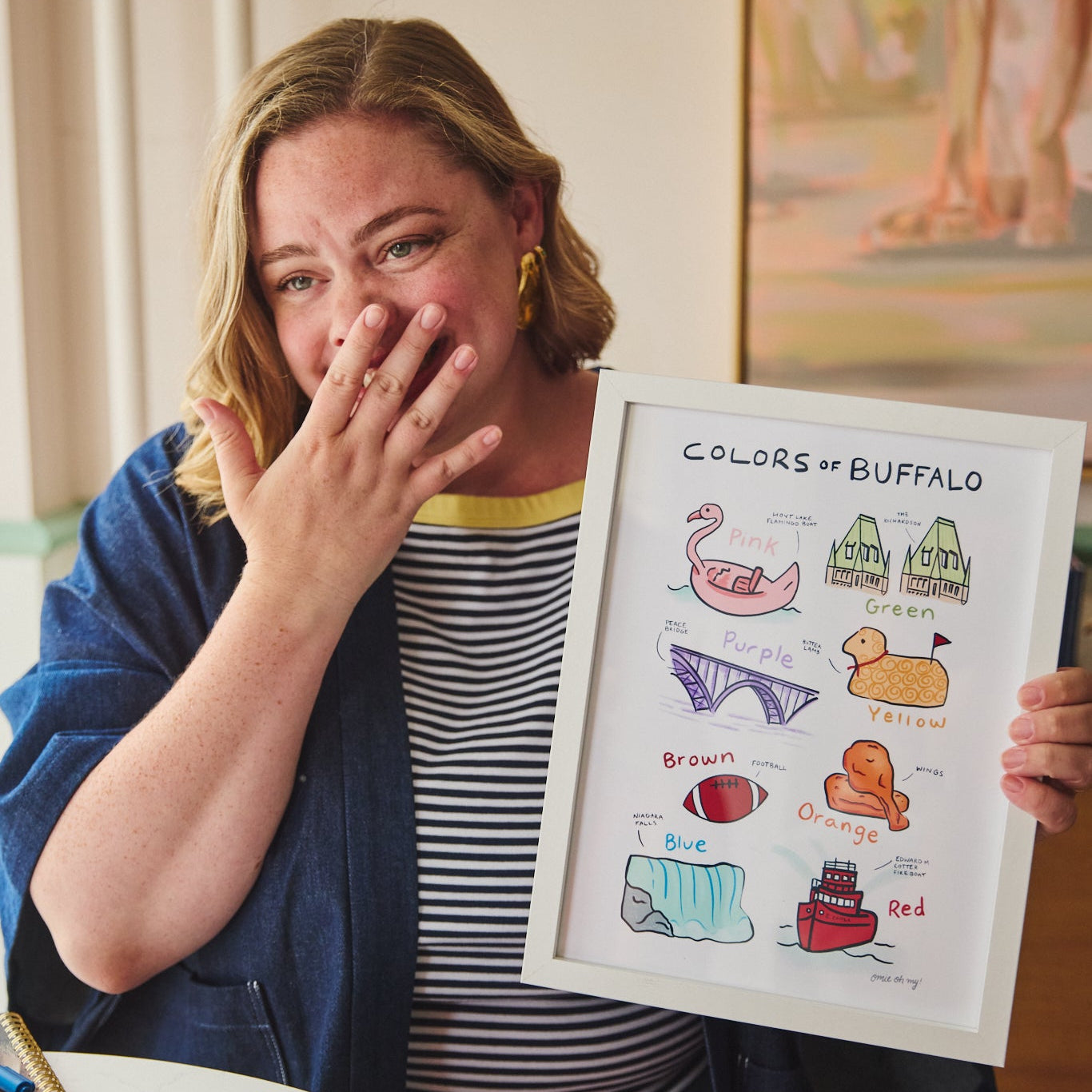 A woman in a blue cardigan and striped shirt holds up a framed art print titled 'COLORS OF BUFFALO' showing various illustrated colors and objects. The setting is indoors with a painting in the background.