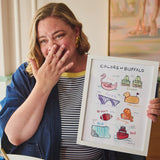 A woman in a blue cardigan and striped shirt holds up a framed art print titled 'COLORS OF BUFFALO' showing various illustrated colors and objects. The setting is indoors with a painting in the background.