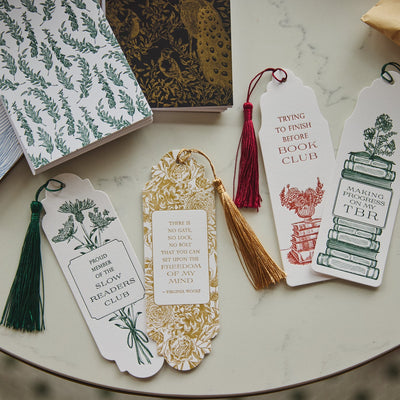 A styled flat-lay of decorative paper bookmarks with tassels arranged on a round marble table, alongside patterned book covers and a kraft envelope.