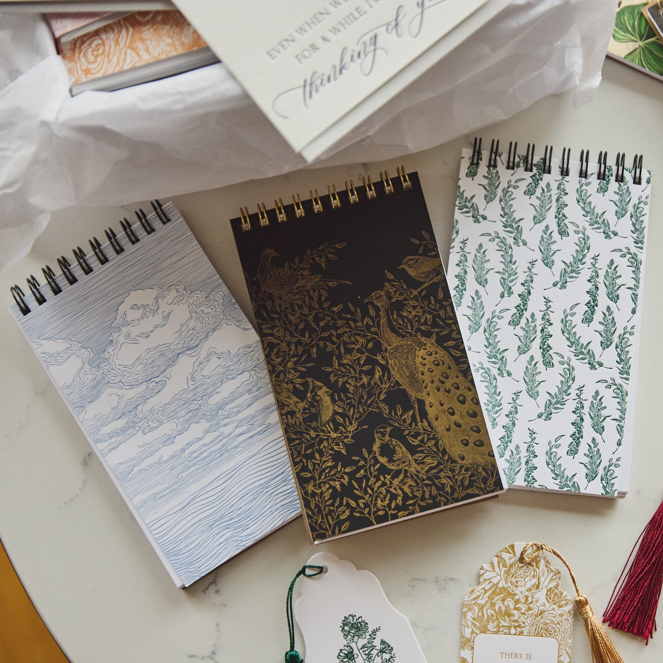 Three spiral-bound notebooks with botanical and animal illustrations are arranged on a light marble surface alongside paper tags, wrapping paper, and a notebook.
