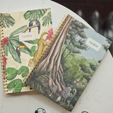 Two spiral-bound notebooks with tropical and forest illustrations resting on a white marble tabletop. One notebook features a beige cover with green leaves, colorful birds, a toucan, and a leopard, while the other has a green forest scene with trees and mushrooms.
