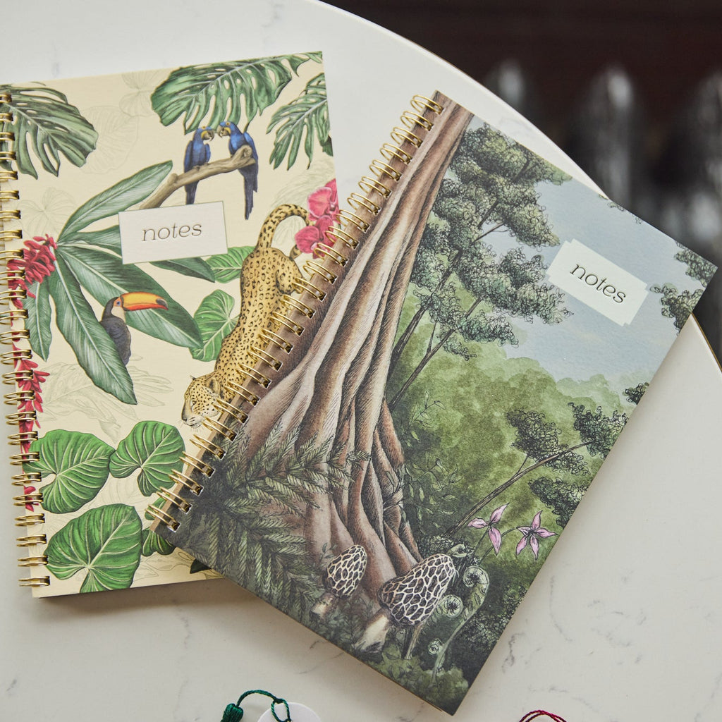 Two spiral-bound notebooks with tropical and forest illustrations resting on a white marble tabletop. One notebook features a beige cover with green leaves, colorful birds, a toucan, and a leopard, while the other has a green forest scene with trees and mushrooms.