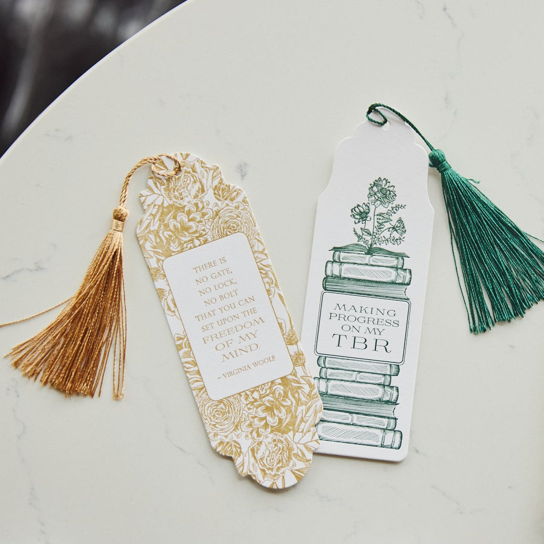 A styled flat-lay of two decorative bookmarks on a white marble surface next to notebooks. The bookmarks include one with a gold foil pattern and a gold tassel, and another with a green tassel.