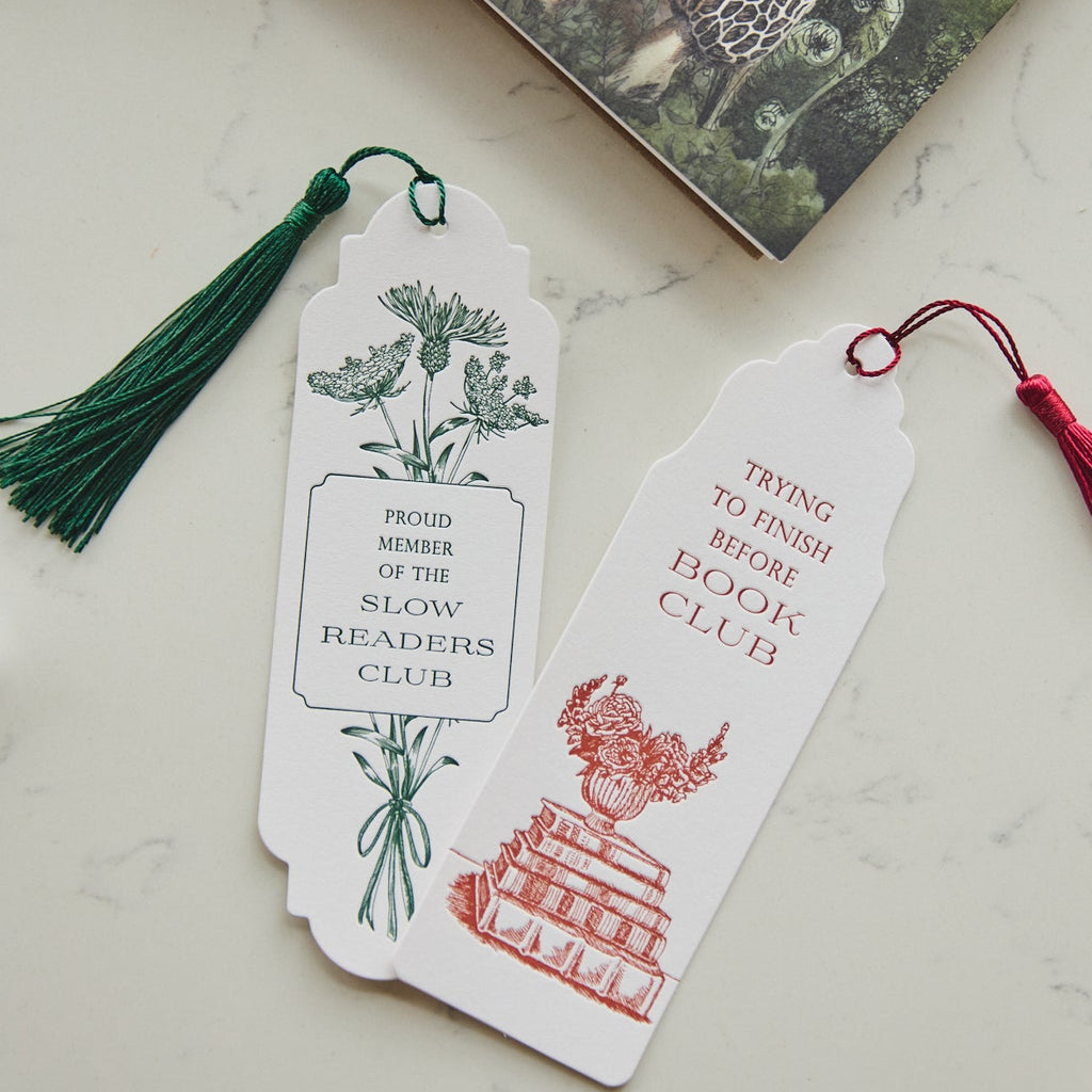 Two decorative paper bookmarks with tassels resting on a white marble surface. One bookmark is white with a green botanical illustration and green tassel, reading 'PROUD MEMBER OF THE SLOW READER'S CLUB.
