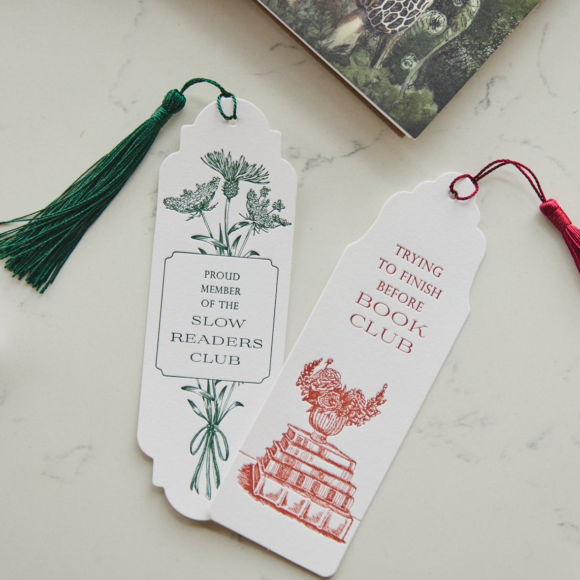 Two decorative paper bookmarks with tassels resting on a white marble surface. One bookmark is white with a green botanical illustration and green tassel, reading 'PROUD MEMBER OF THE SLOW READER'S CLUB.