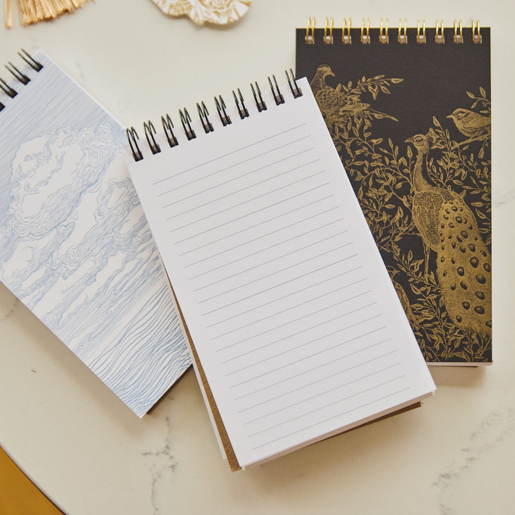 Three spiral-bound notebooks arranged on a light marble surface. One notebook has a white page with light blue horizontal lines, one is closed with a black cover featuring gold peacock and floral artwork, and one is partially visible with a pale blue patterned cover.