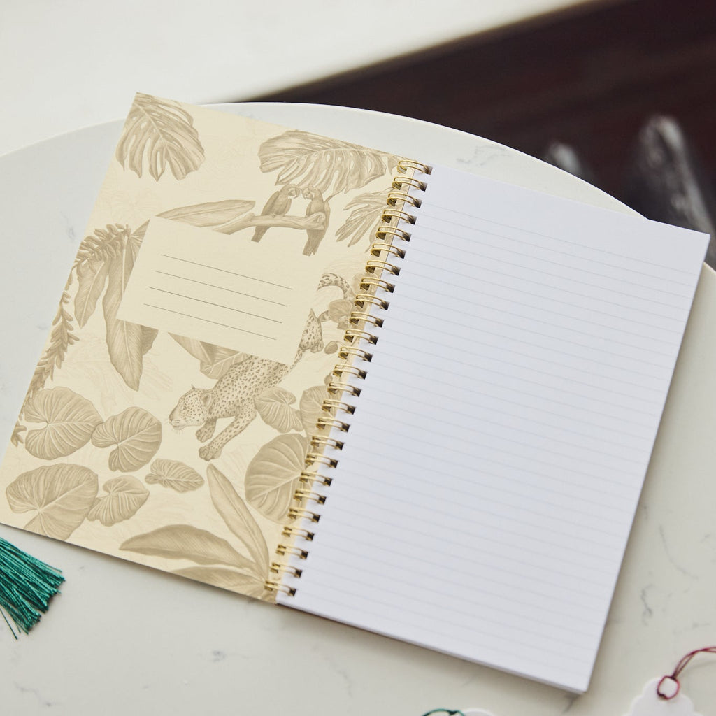 A spiral-bound notebook with a beige botanical patterned cover and white lined pages, resting on a round white table. A green tassel tag and a small card are partially visible nearby.