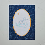 A square art print featuring a white duck swimming in a light blue pond, framed by a scalloped gold oval border on a dark navy blue background with starry speckles and faint duck silhouettes. The print is photographed flat against a light gray marble surface.