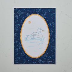 A square art print featuring a white duck swimming in a light blue pond, framed by a scalloped gold oval border on a dark navy blue background with starry speckles and faint duck silhouettes. The print is photographed flat against a light gray marble surface.