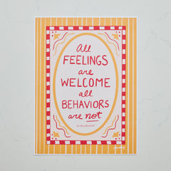 A square art print with a colorful quote reading 'All Feelings are Welcome, all Behaviors are not'. The print features a red and white checkered border, a yellow oval frame, and a yellow and white striped background, displayed on a light gray marble surface.