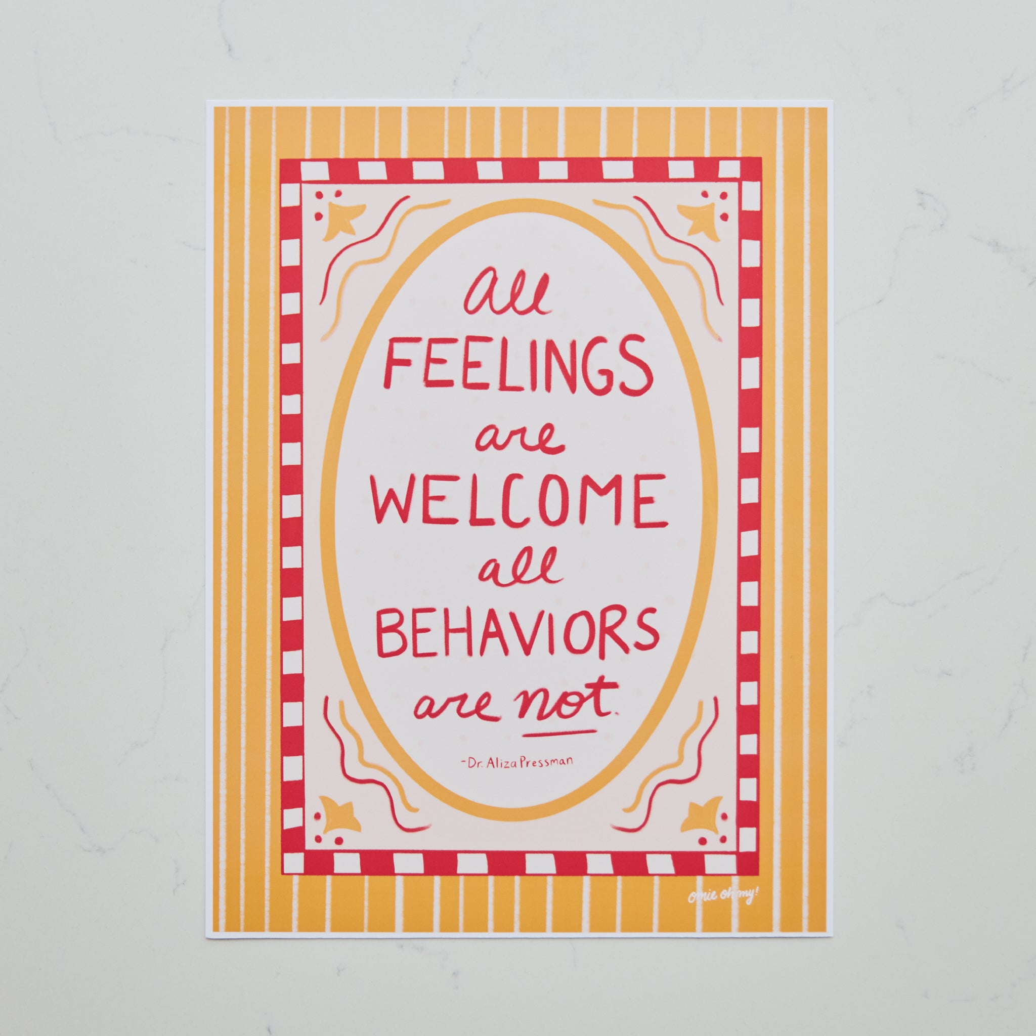 A square art print with a colorful quote reading 'All Feelings are Welcome, all Behaviors are not'. The print features a red and white checkered border, a yellow oval frame, and a yellow and white striped background, displayed on a light gray marble surface.