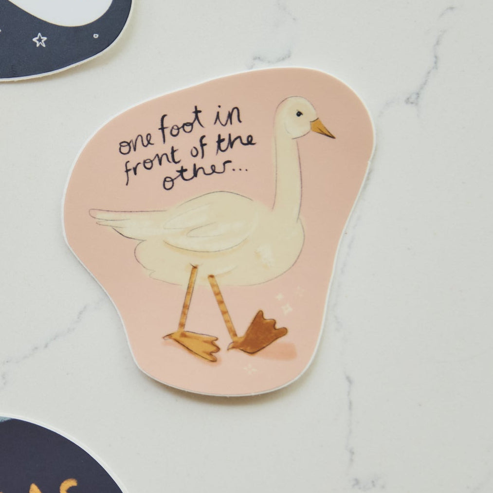 A die-cut sticker featuring a white goose illustration and the handwritten text 'one foot in front of the other.