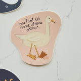 A die-cut sticker featuring a white goose illustration and the handwritten text 'one foot in front of the other.