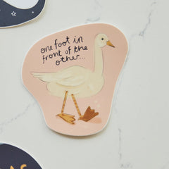A die-cut sticker featuring a white goose illustration and the handwritten text 'one foot in front of the other.