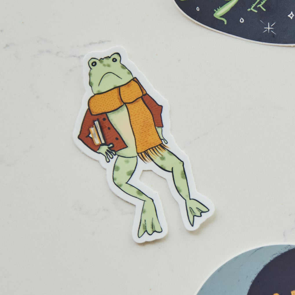 A die-cut sticker featuring a green frog wearing an orange scarf and a brown jacket, holding a book.