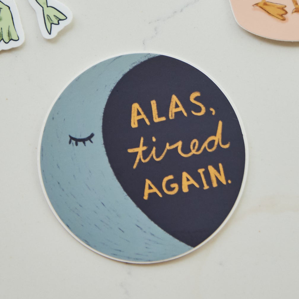 A round sticker with a crescent moon design, featuring the text 'ALAS, tired AGAIN.' in gold lettering on a dark navy and light blue background, placed on a light marble surface.
