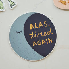 A round sticker with a crescent moon design, featuring the text 'ALAS, tired AGAIN.' in gold lettering on a dark navy and light blue background, placed on a light marble surface.