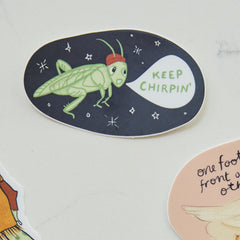 An oval sticker featuring a green grasshopper wearing a red cap with a speech bubble that says 'KEEP CHIRPIN'', shown on a light surface next to other partial stickers.