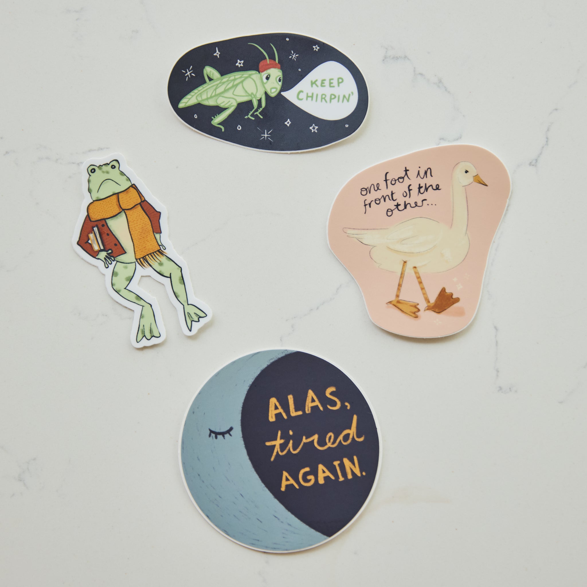 Four whimsical die-cut stickers arranged on a light marble surface. The stickers feature illustrated frogs, a goose, and text-based designs in pastel and dark colors.