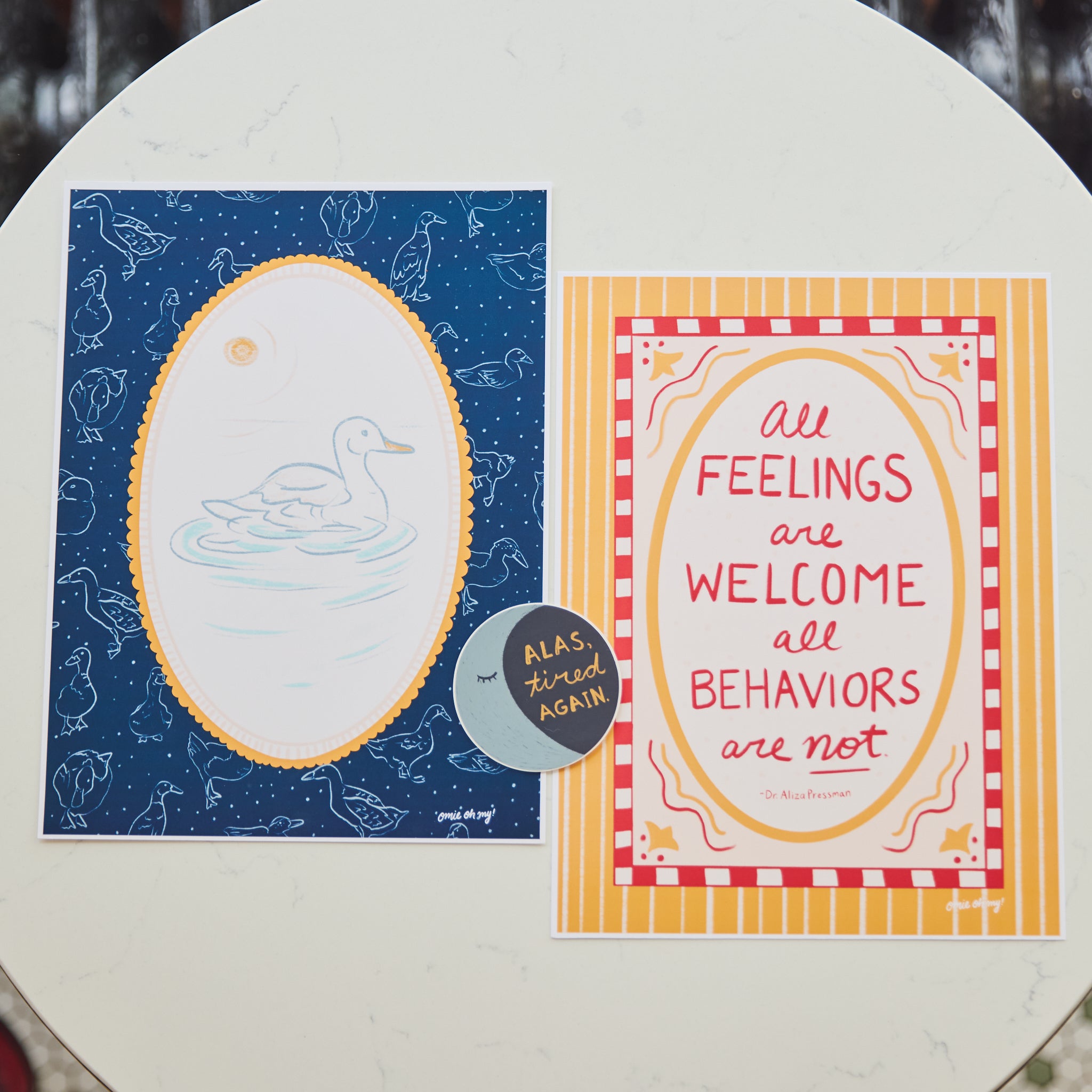Two printed cards placed side by side on a round white table. The left card has a dark blue background with a white duck illustration inside a yellow oval border. The right card has the text "all feelings are welcome all behaviors are not" in red on a yellow and pink background.