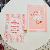 Two pink and white printed cards or art prints displayed on a round white table. One card features the text 'All feelings are welcome all behaviors are not' in red lettering inside an ornate border, and the other shows a white goose with one foot in front of its own reflection on a pink striped background.