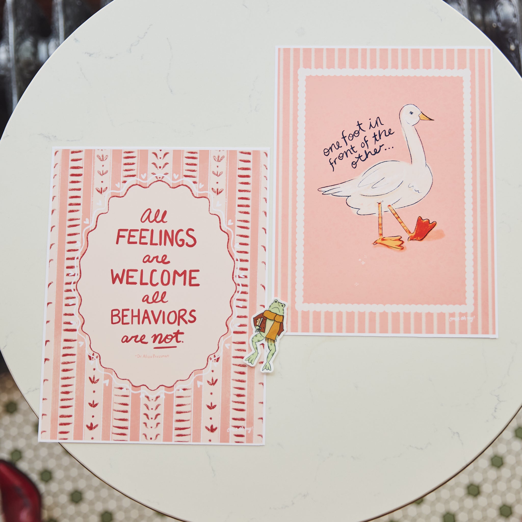 Two pink and white printed cards or art prints displayed on a round white table. One card features the text 'All feelings are welcome all behaviors are not' in red lettering inside an ornate border, and the other shows a white goose with one foot in front of its own reflection on a pink striped background.