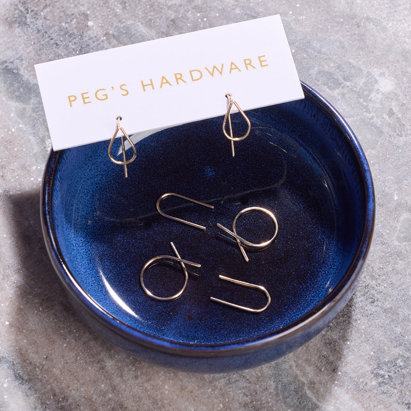Gold-toned geometric dangle earrings displayed in a small dark blue ceramic dish, with a white card reading 'PEG'S HARDWARE' resting above them on a gray stone surface.