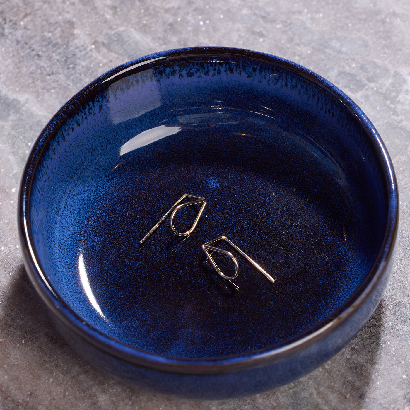 A pair of small gold geometric hoop earrings displayed inside a deep blue glazed ceramic bowl on a gray stone surface.