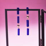 Two of Peg's Hardware's Capri earrings, hanging from a black metal frame.