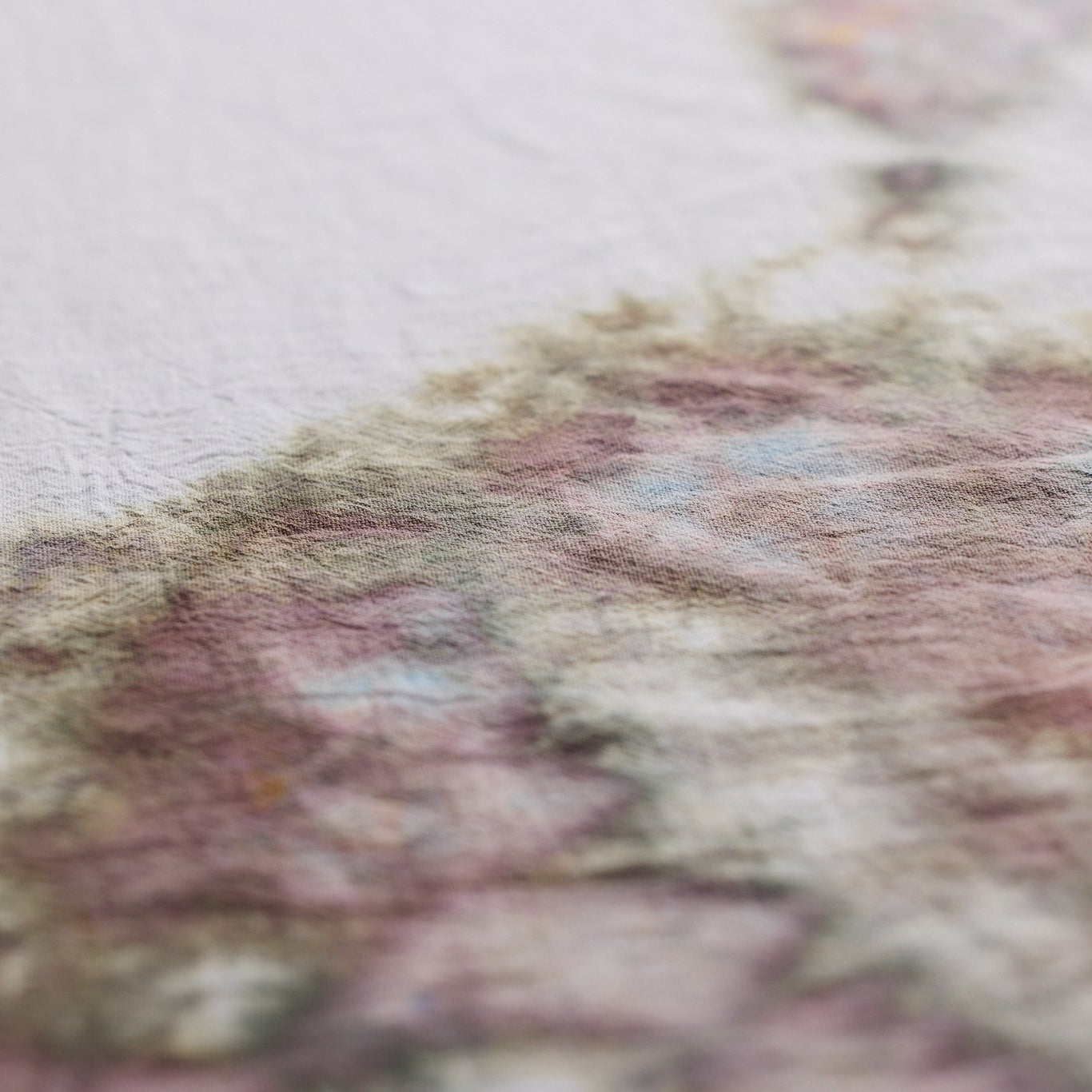A close-up of a textured dyed fabric showing a soft, painterly transition from off-white on the left to dusty pink, mauve, and muted green on the right.