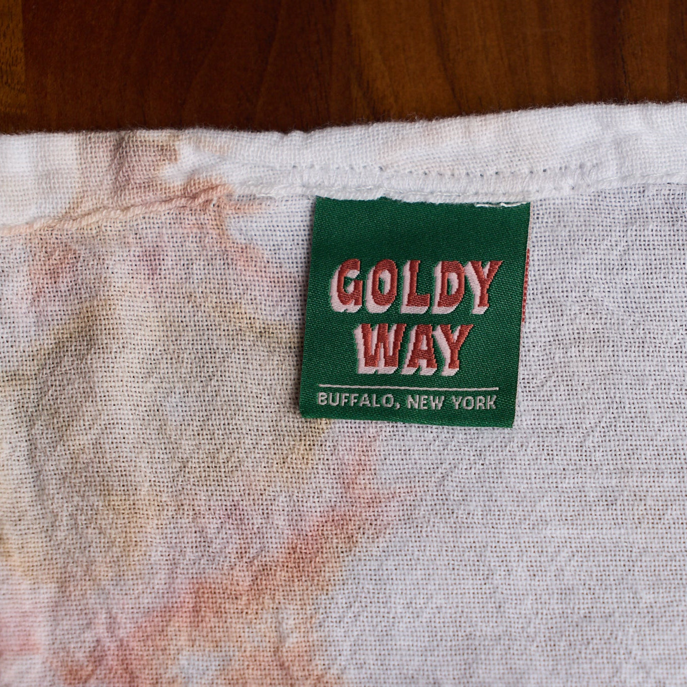 Close-up of a vintage Goldy Way t-shirt neck label on a white cotton fabric with pink and orange tie-dye stains, set against a dark wood background.