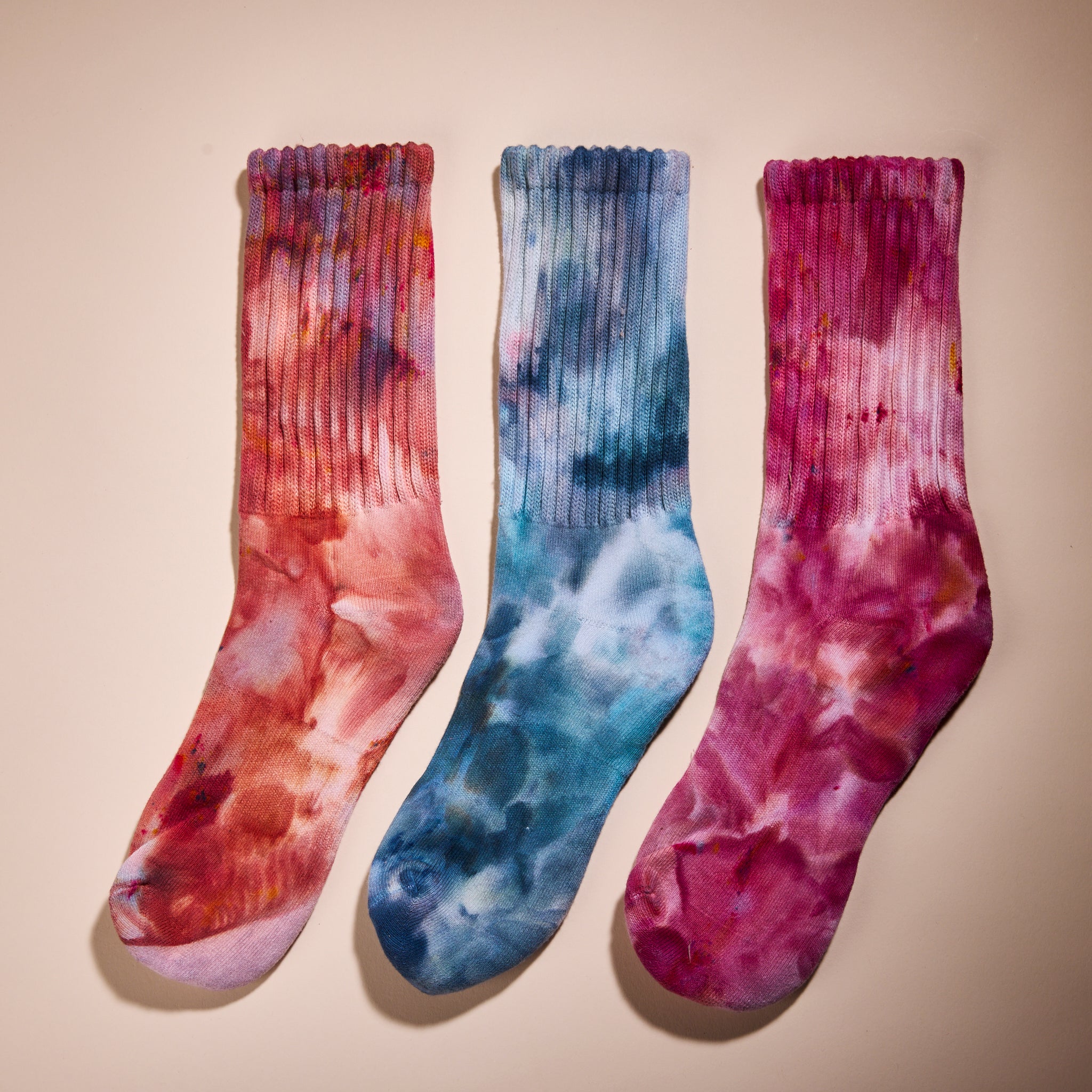 three of Goldy Way's hand-dyed crew socks, each in a different color. From left to right, colors shown are Solstice, (orange with reds and purple), Lake Effect (blue with darker blue and pink), and Winterberry (dark fuchsia with blue and orange).