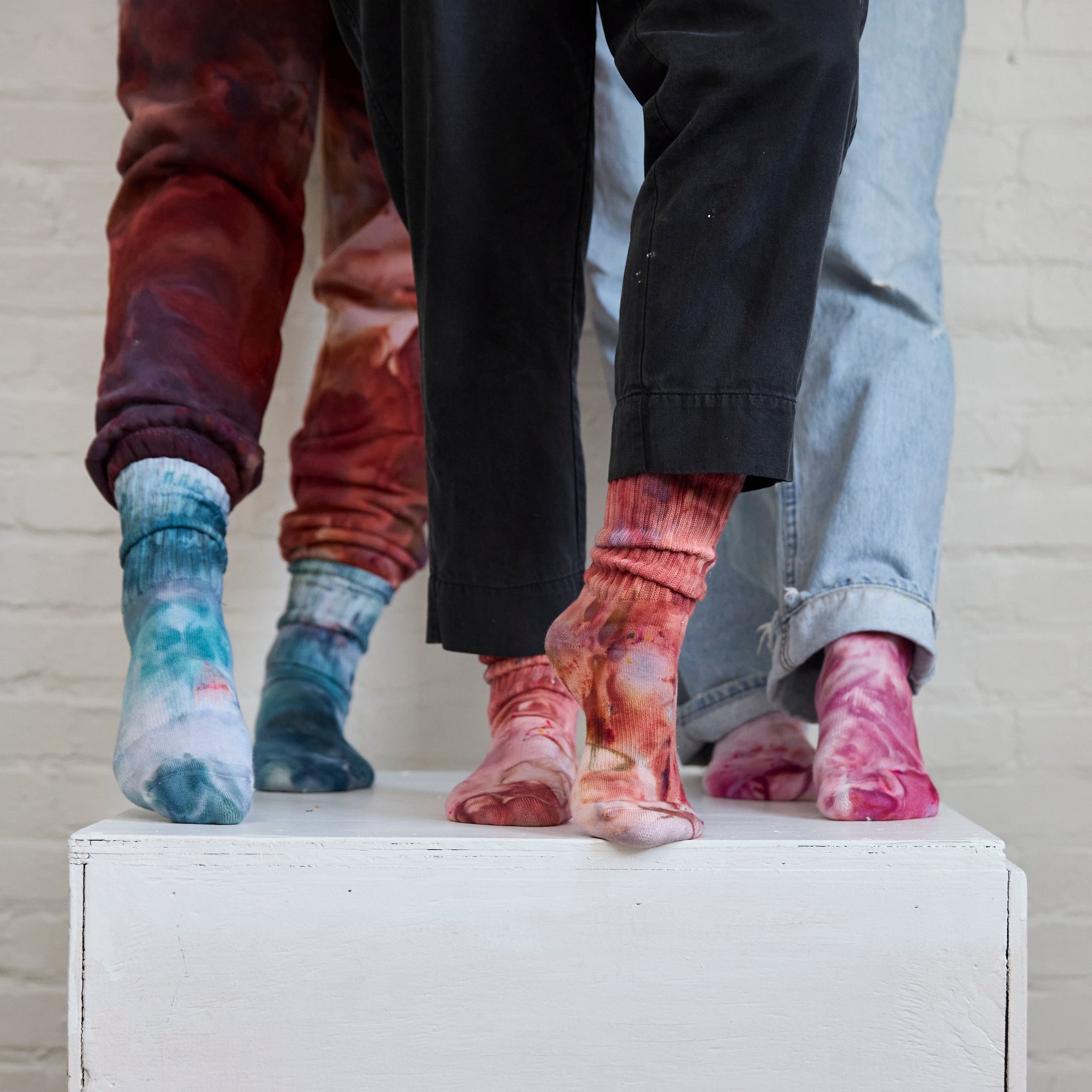 Three pairs of Goldy Way's hand-dyed crew socks, each pair on the feet of a model. From left to right, colors shown are Lake Effect (blue with darker blue and pink), Solstice (orange with reds and purple), and Winterberry (dark fuchsia with blue and orange).
